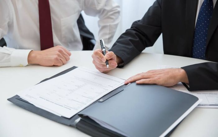 person in black suit preparing to sign a document next to person in red tie